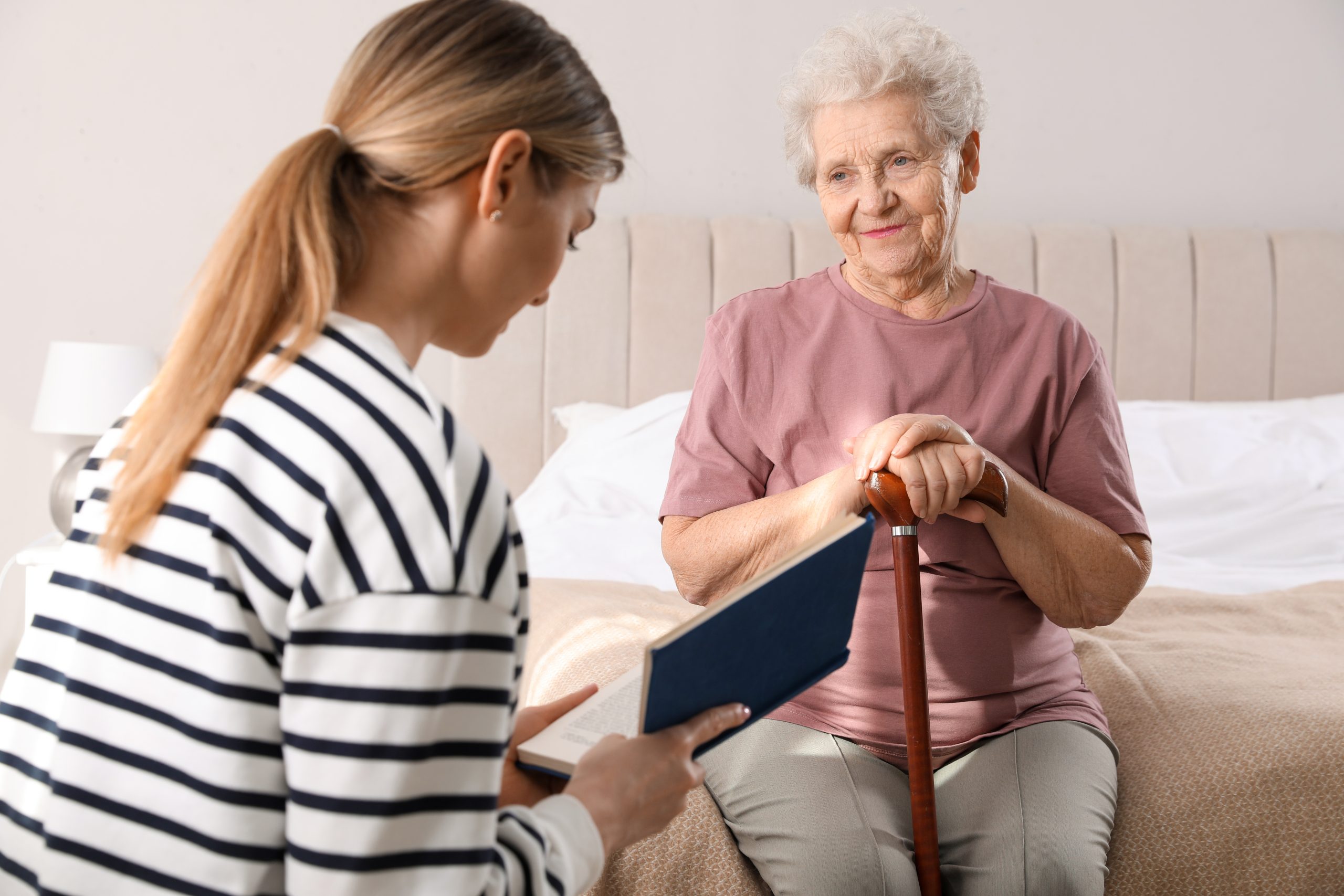 Young Caregiver Reading Book To Senior Woman In Bedroom. Home Ca